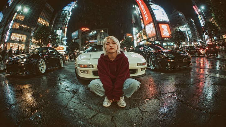 Potret olahan AI: Sosok ber-hoodie burgundi berjongkok di depan mobil pucat di jalan malam yang berkilau oleh lampu kota. Komposisi horizontal.